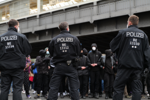 Eine Gruppe von Polizisten in Uniform steht vor einer Menge von Menschen, die ebenfalls schwarze Uniformen und Masken tragen, mit einer Brücke und einem Gebäude im Hintergrund, was auf eine städtische Umgebung hindeutet.