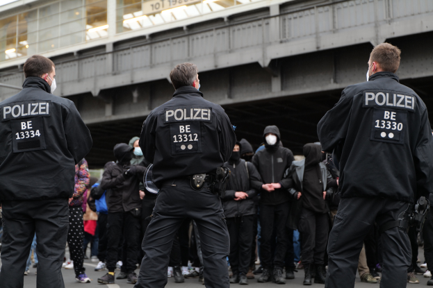 Eine Gruppe von Polizisten in Uniform steht vor einer Menge von Menschen, die ebenfalls schwarze Uniformen und Masken tragen, mit einer Brücke und einem Gebäude im Hintergrund, was auf eine städtische Umgebung hindeutet.