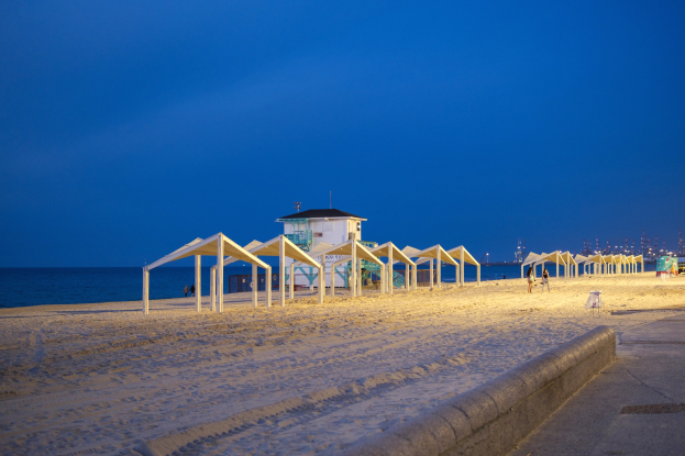 Ein Strand bei Nacht mit einem Rettungsturm, Menschen am Ufer, einem Zaun, Zelten, einem Haus, Pfählen, Wasser, Lichtern und einem bewölkten Himmel.