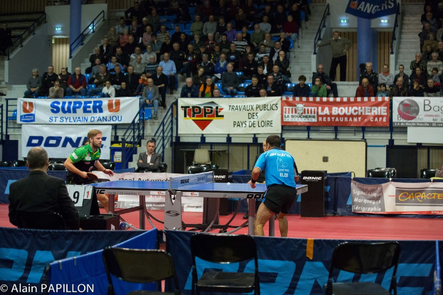 Zwei Männer spielen drinnen Tischtennis, mit mehreren Zuschauern, die stehen und sitzen, einigen leeren Stühlen und Werbeplakaten, mit Text in der unteren linken Ecke.