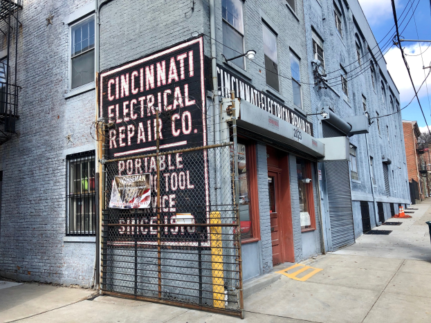 Außenansicht des Cincinnati Electrical Repair Co. Gebäudes mit Beschilderung, einem Metallzaun, einem Müllcontainer, Strommasten und -kabeln, Bäumen und einem bewölkten Himmel.