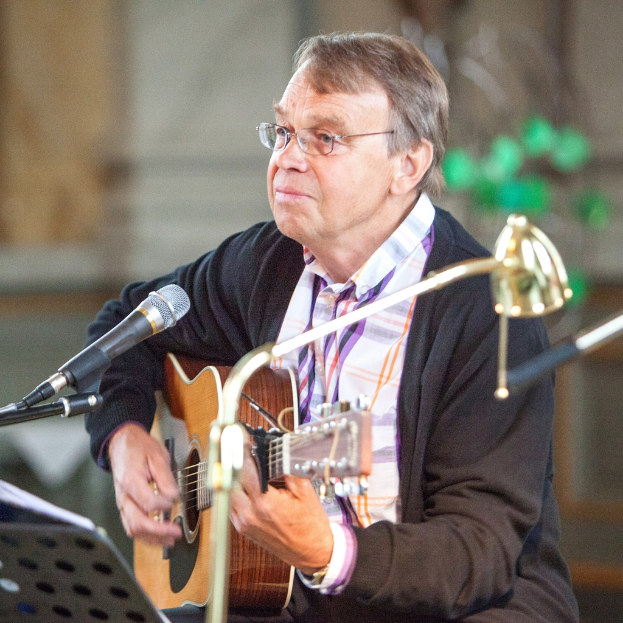 Ein Mann in einer schwarzen Jacke spielt Gitarre und trägt eine Brille, vor ihm steht ein Mikrofon.