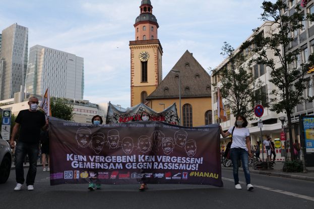 Eine Gruppe von Menschen in Masken, die eine Straße entlanggehen und ein Banner halten, mit einem geparkten Auto auf der linken Seite, Gebäuden und Bäumen im Hintergrund und einem Uhrenturm unter einem klaren blauen Himmel.