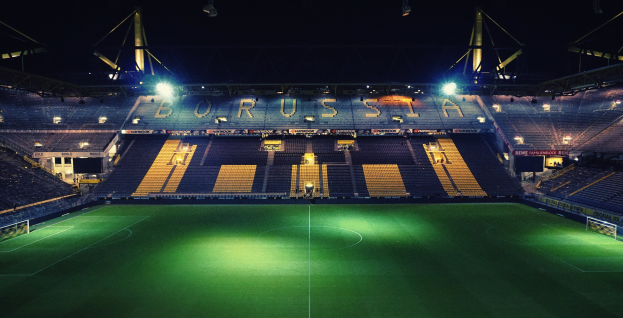 Ein Fußballfeld bei Nacht, beleuchtet von hellen Stadionlampen, mit einigen Zuschauern auf den Tribünen und saftig grünem Rasen.