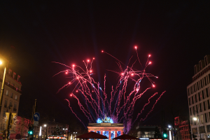 Eine belebte Stadtstraße bei Nacht mit vielen Menschen, Fahrzeugen, Zelten und Gebäuden sowie einem farbenfrohen Feuerwerk am Himmel im Hintergrund.