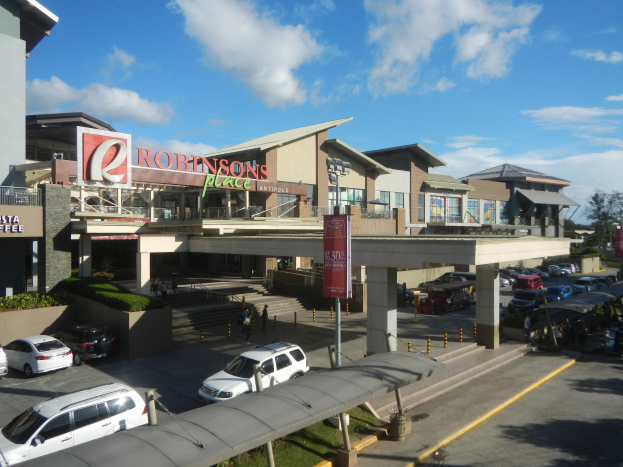 Ein belebtes Einkaufszentrum mit Gebäuden, Geschäften, Fahrzeugen, Fußgängern, Straßenmöblierung, Grünflächen und einer bewölkten Himmel.