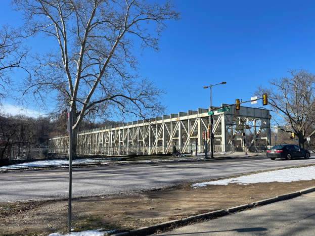 Ein Auto fährt auf einer schneebedeckten Straße neben einer Brücke, mit Laternen, Ampeln, Bäumen und einem bewölkten Himmel im Hintergrund.